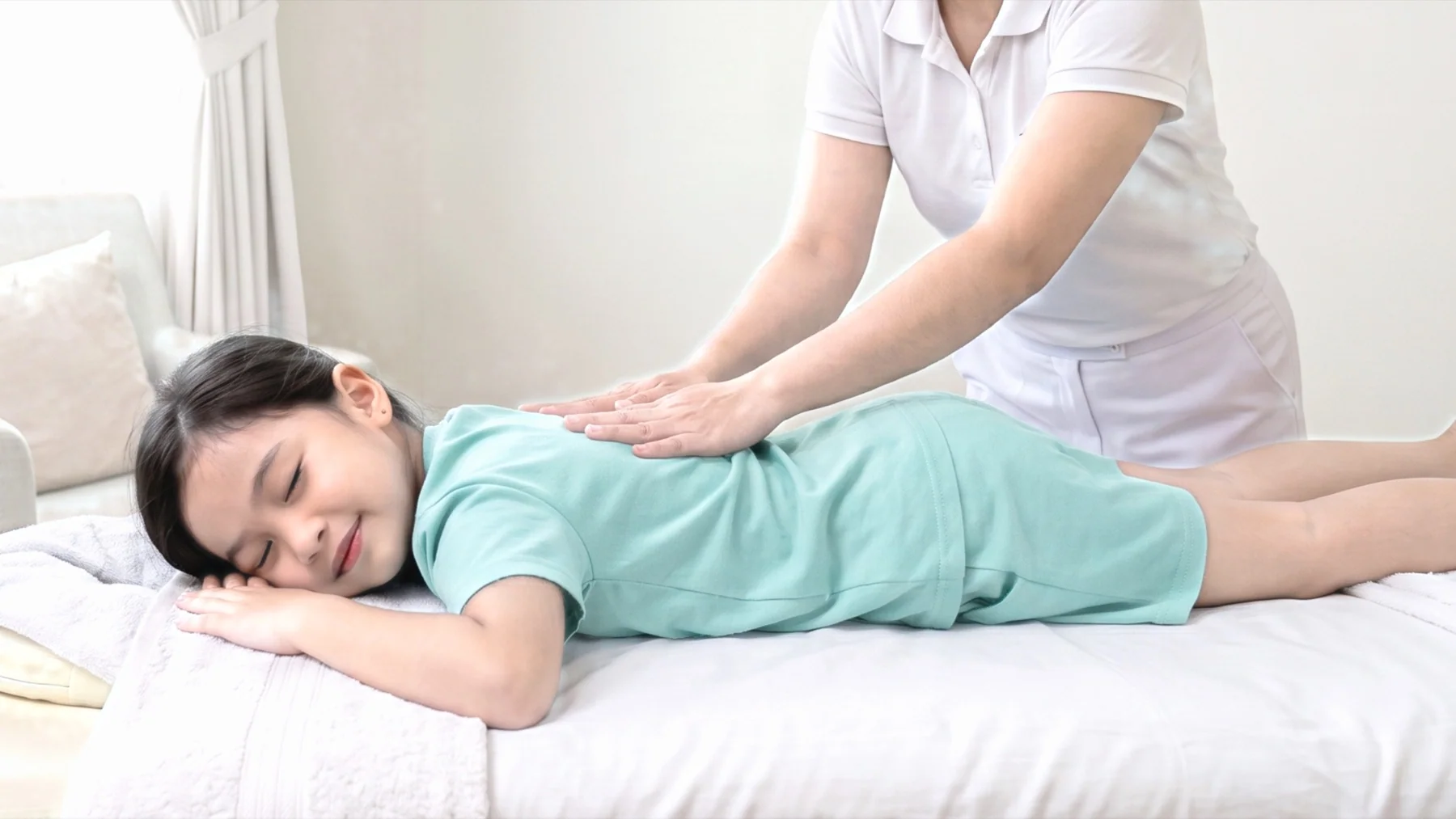 A child sitting comfortably with a parent nearby while a professional therapist explains a gentle, age-appropriate hand or shoulder massage. Fully clothed, welcoming, authentic home setting.